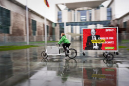 Vor dem Kanzleramt in Berlin: Eine Greenpeace-Aktivistin fährt mit dem Lastenrad und Anhänger mit einem Schild: Ein Bild von Olaf Scholz im Stil eines Walplakates, daneben "Kanzler für Verbrennerschutz"