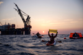 Vor Borkum auf dem Meer. Im Hintergrund ist die Plattform-Sleipnir, davor protestieren Greenpeace-Aktivist:innen gegen die Gas-Bohrungen: Zwei Kajaks und eine Rettungsinsel, ein Kajakee hält ein Schild: Geen nieuw Gas