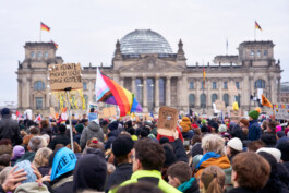 Während einer Demonstration: Blick auf die Menschenmenge und den Reichstag in Berlin. Im Vordergrund ist eine LGBTQ-Flagge und ein Schild: "Ich könnte mich um schönere Dinge kümmern".