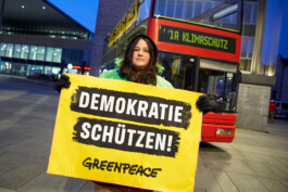 Eine Greenpeace-Aktivistin hält ein Banner "Demokratie schützen!" in die Kamera, im Hintergrund ist ein Bus mit der Aufschrift "1A Klimaschutz".