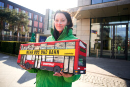 Eine Greenpeace-Aktivistin hält das Modell eines Busses in die Kamera, auf dem Bus stehtn "Mehr Bus und Bahn". In dem Bus sind Unterschriften, die übergeben werden sollen.