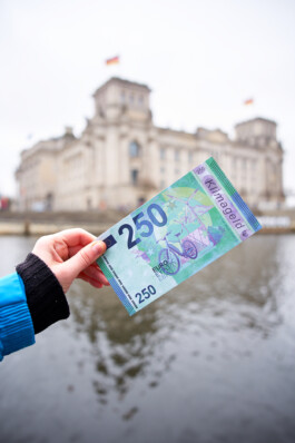 Ein 250-€-Schein mit dem Aufdruck "Klimageld" wird in die Kamera gehalten, im Hintergrund ist der Reichstag in Berlin.