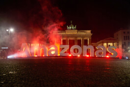 Vor dem Brandenburger Tor in Berlin stehen Buchstaben, die das Wort "Amazonas" bilder. Hinter den Buchstaben ist Rauch (Nebelmaschine) und eine Lichtinstallation.