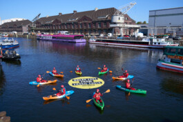 Im Westhafen in Berlin sind mehrere Kajaks um ein rundes, gelbes Schwimmbanner herum, wie Sonnenstrahlen, angeordnet. Auf dem Banner steht: "Break free from fossil fuels".